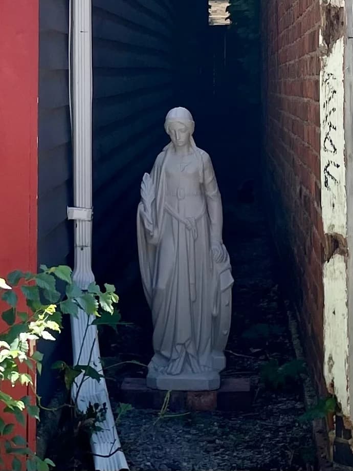 A classical white statue of a robed woman holding a sheaf of wheat, set in a narrow alley between two buildings
