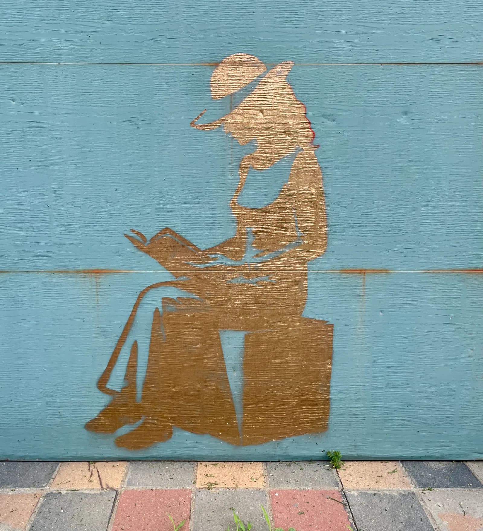 A golden stencil mural of a woman in a wide-brimmed hat, seated and reading a book, painted on a teal wooden wall