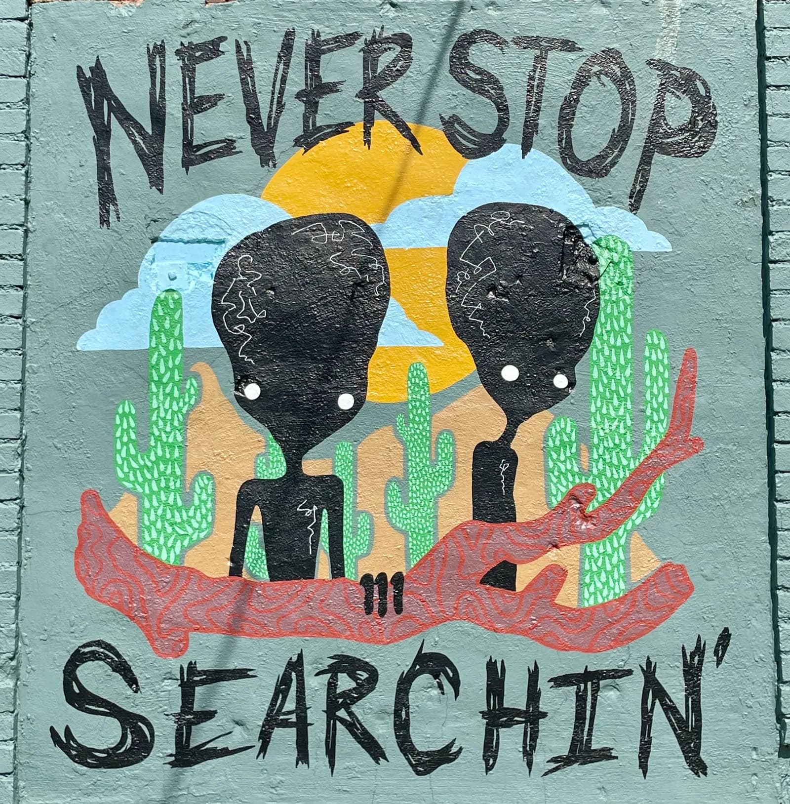 Casey Micksun's “Never Stop Searchin'” mural — two silhouetted figures among cactuses beneath a desert sun, lettered Never Stop Searchin'